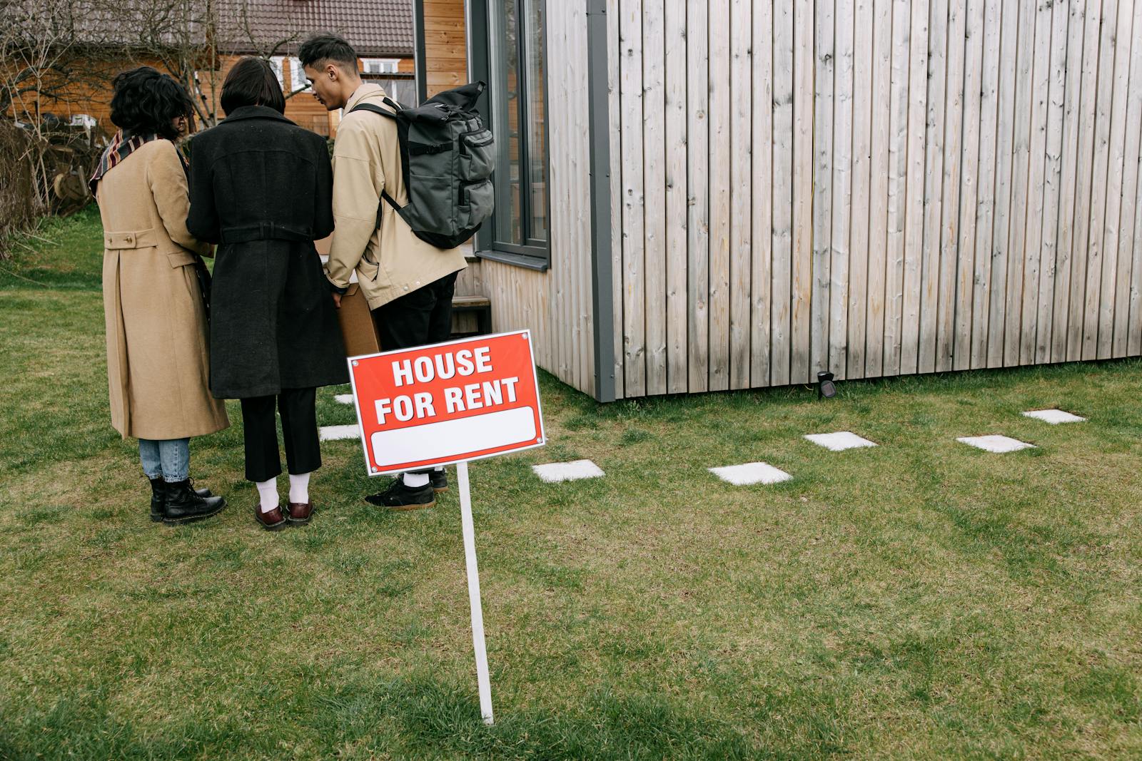 Group of adults viewing a wooden house with a 'House for Rent' sign on the lawn.